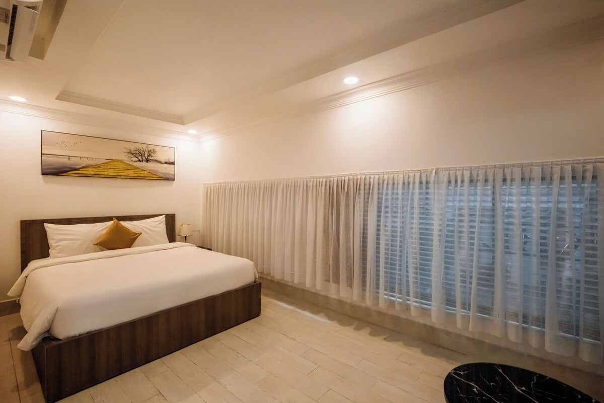 A hotel room with a bed, a painting, a window with curtains, and a view of buildings.