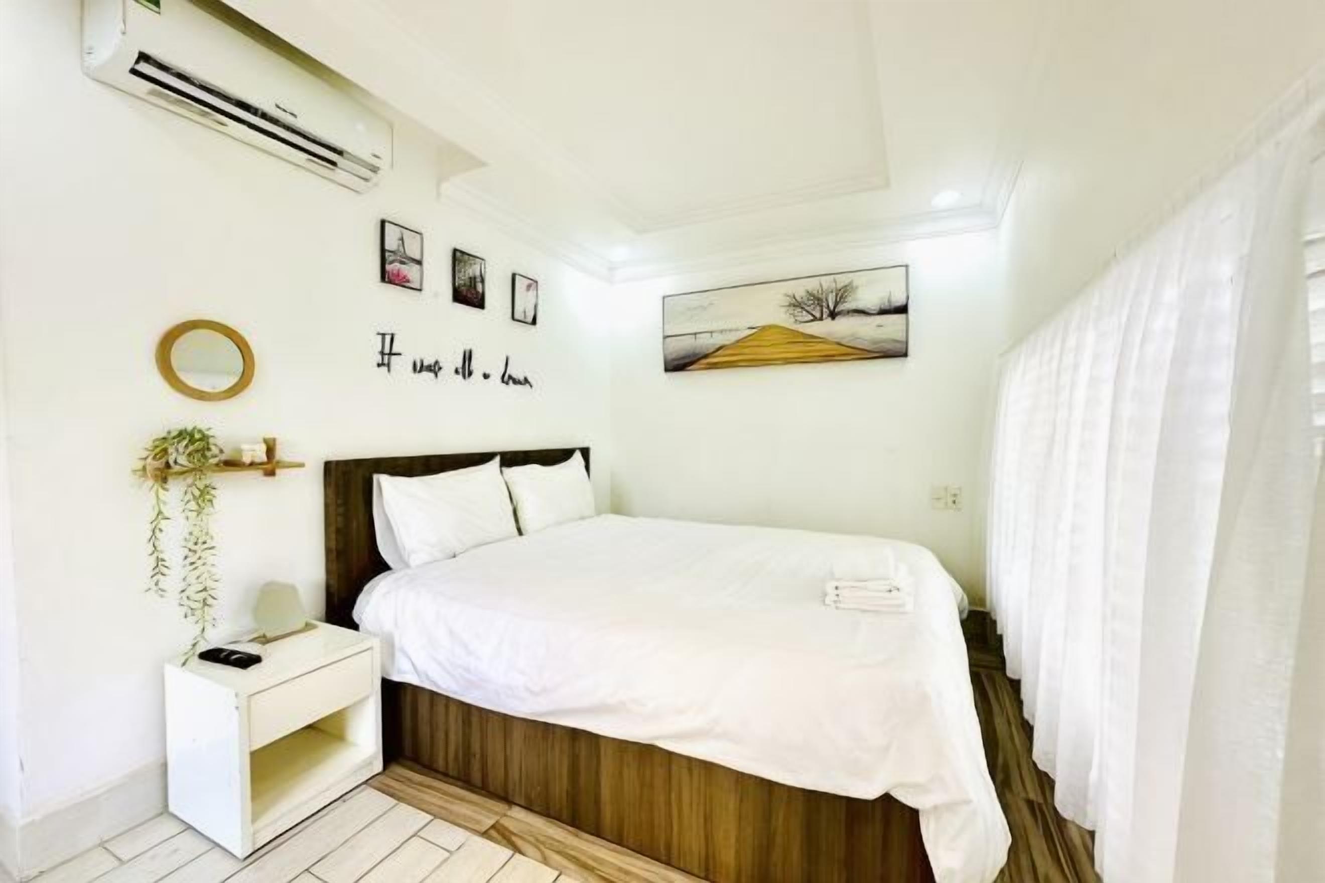 A bedroom with a bed, bedside table, wall-mounted air conditioner, framed pictures, and a painting.