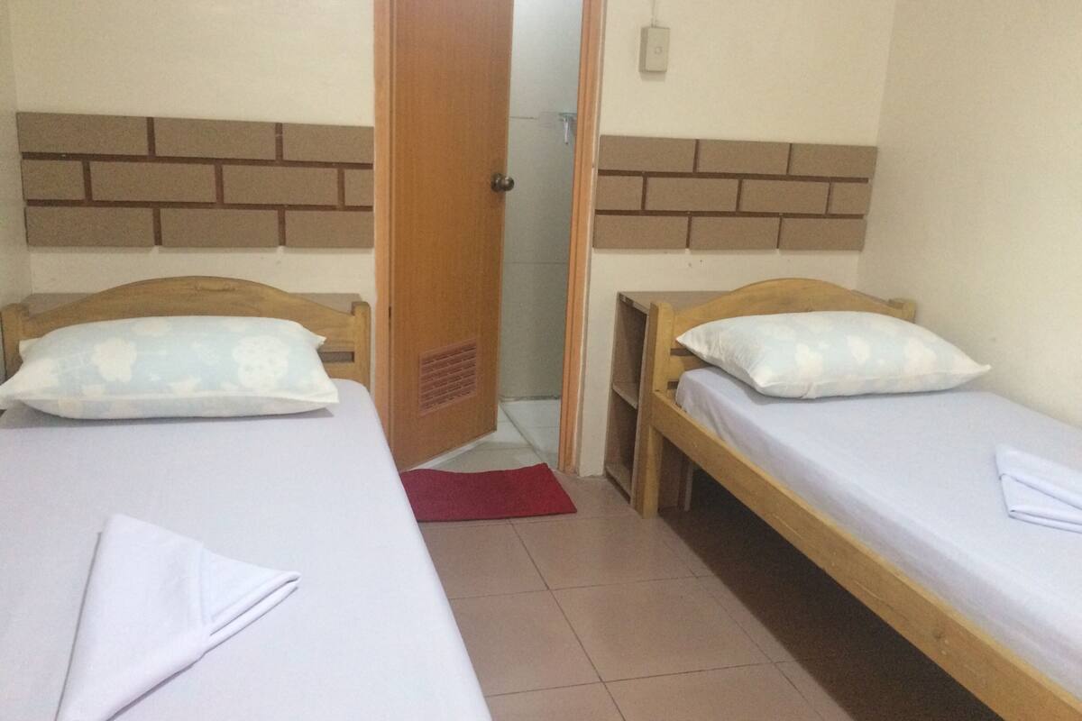 Quarto com duas camas de solteiro, cada uma com cabeceira de madeira e roupa de cama branca. Há um tapete vermelho no chão entre as camas, e uma porta de madeira com uma parede de tijolos ao fundo.