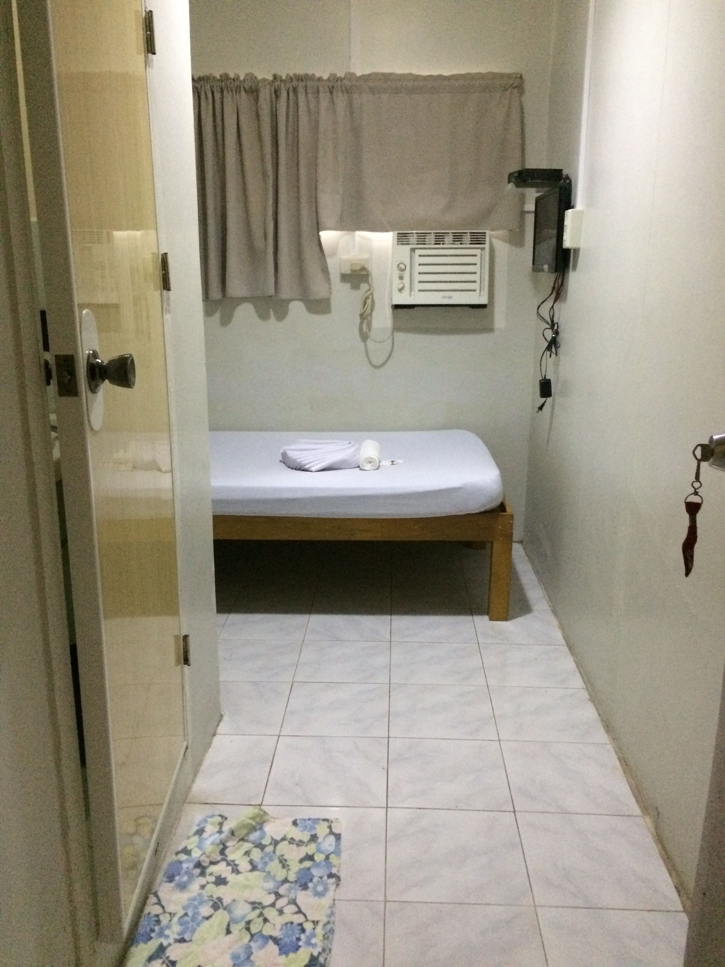 A small, narrow room with a single bed, a wall-mounted heater, and a small rug on the floor.
