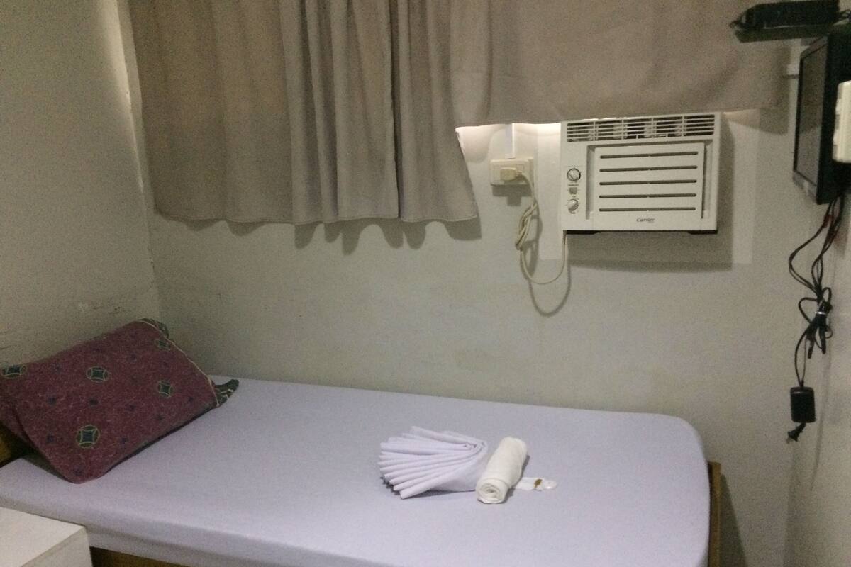 Uma cama de solteiro com colchão branco, travesseiro vermelho e toalha branca. Há um ar condicionado fixado na parede e um telefone.