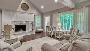 A spacious living room with a fireplace, comfortable seating, and large windows offering a view of the outdoors. - The Farm House (Blythewood)