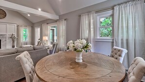 A living room with a round wooden table, a grey sofa, and a white vase with flowers. - The Farm House (Blythewood)