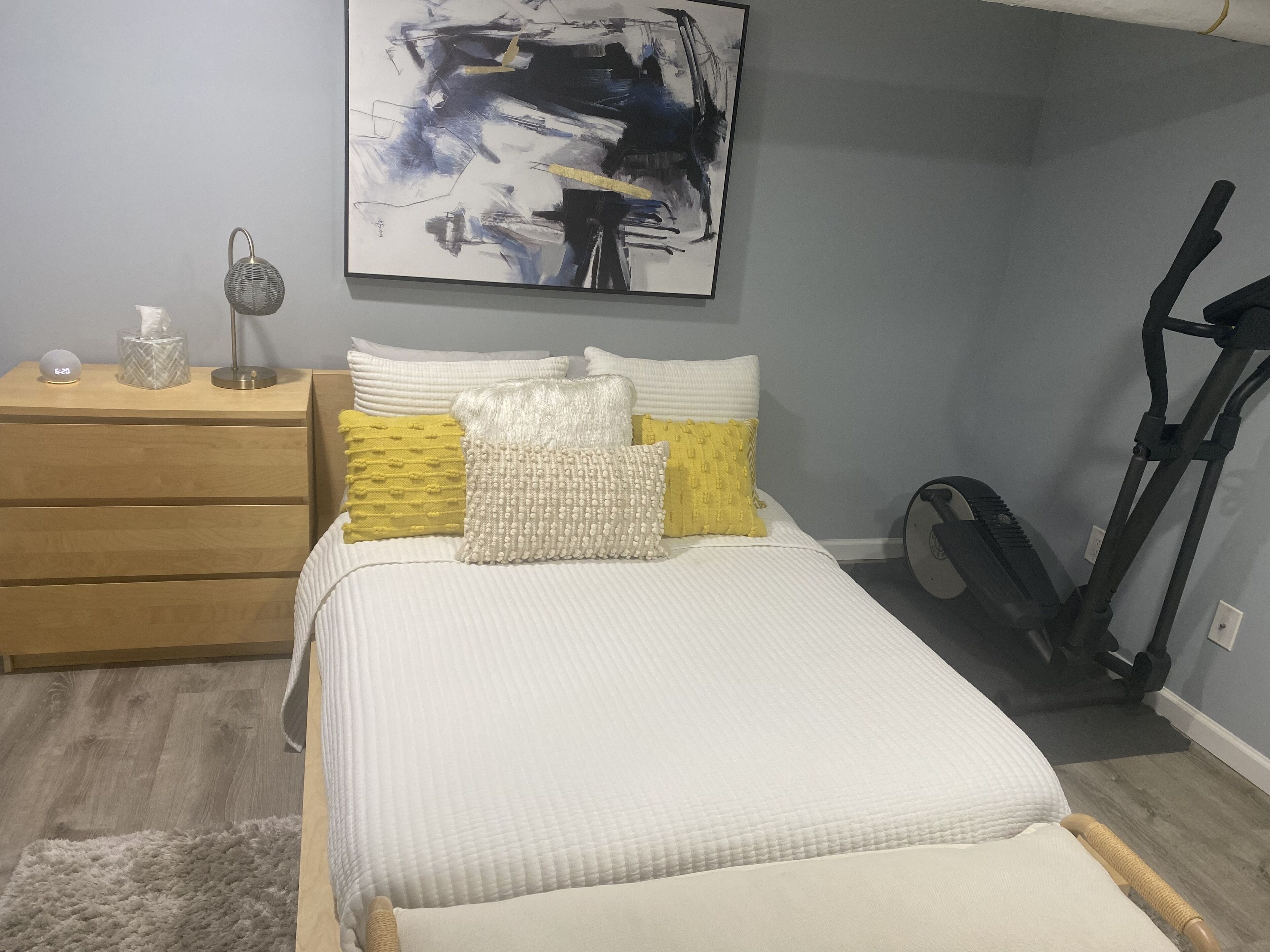 Ein Schlafzimmer mit einem Bett, einem Nachttisch, einer Lampe, einem an der Wand befestigten abstrakten Gemälde und einem stationären Heimtrainer.