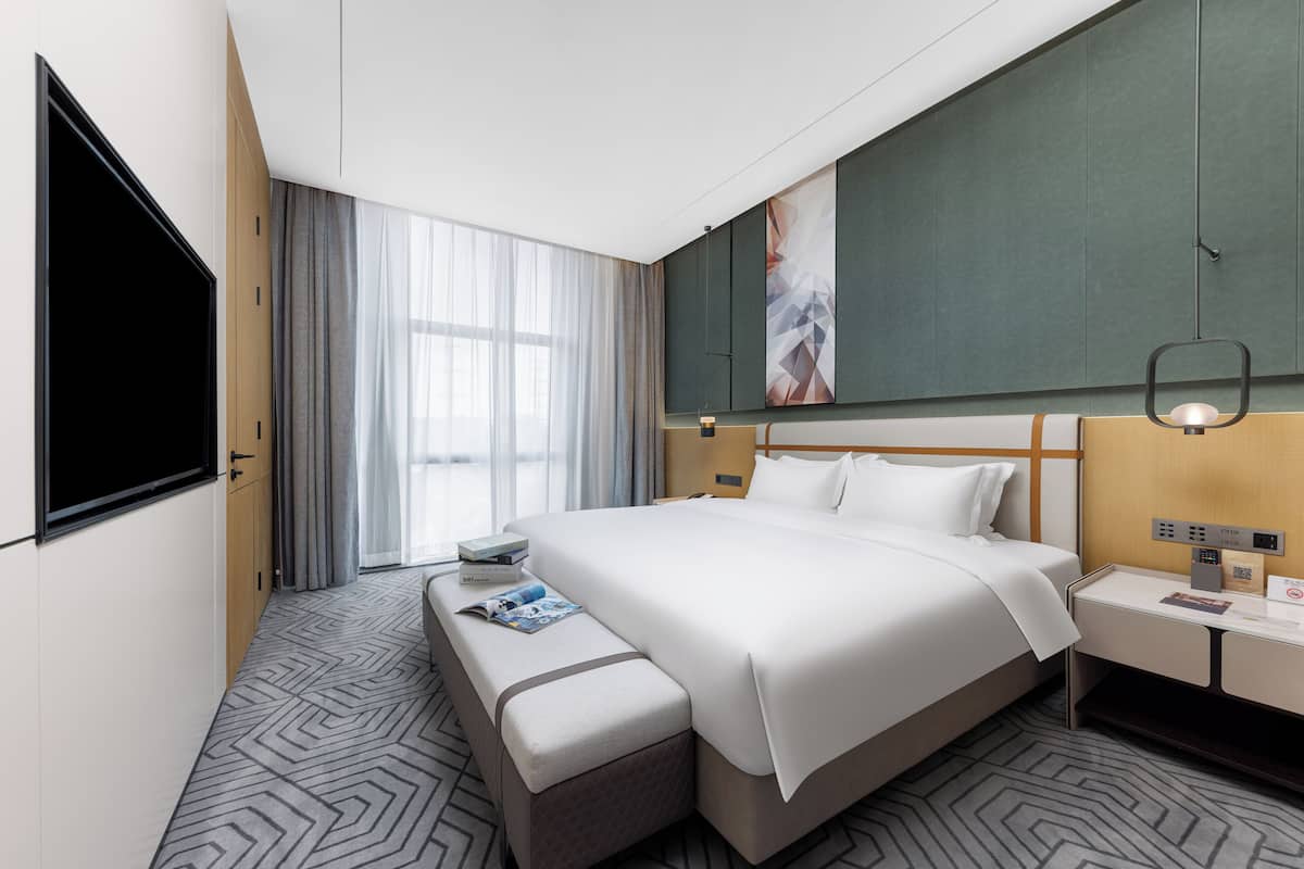 A hotel room with a large bed, a bedside table, a bench with magazines, a TV, and a large window with curtains.