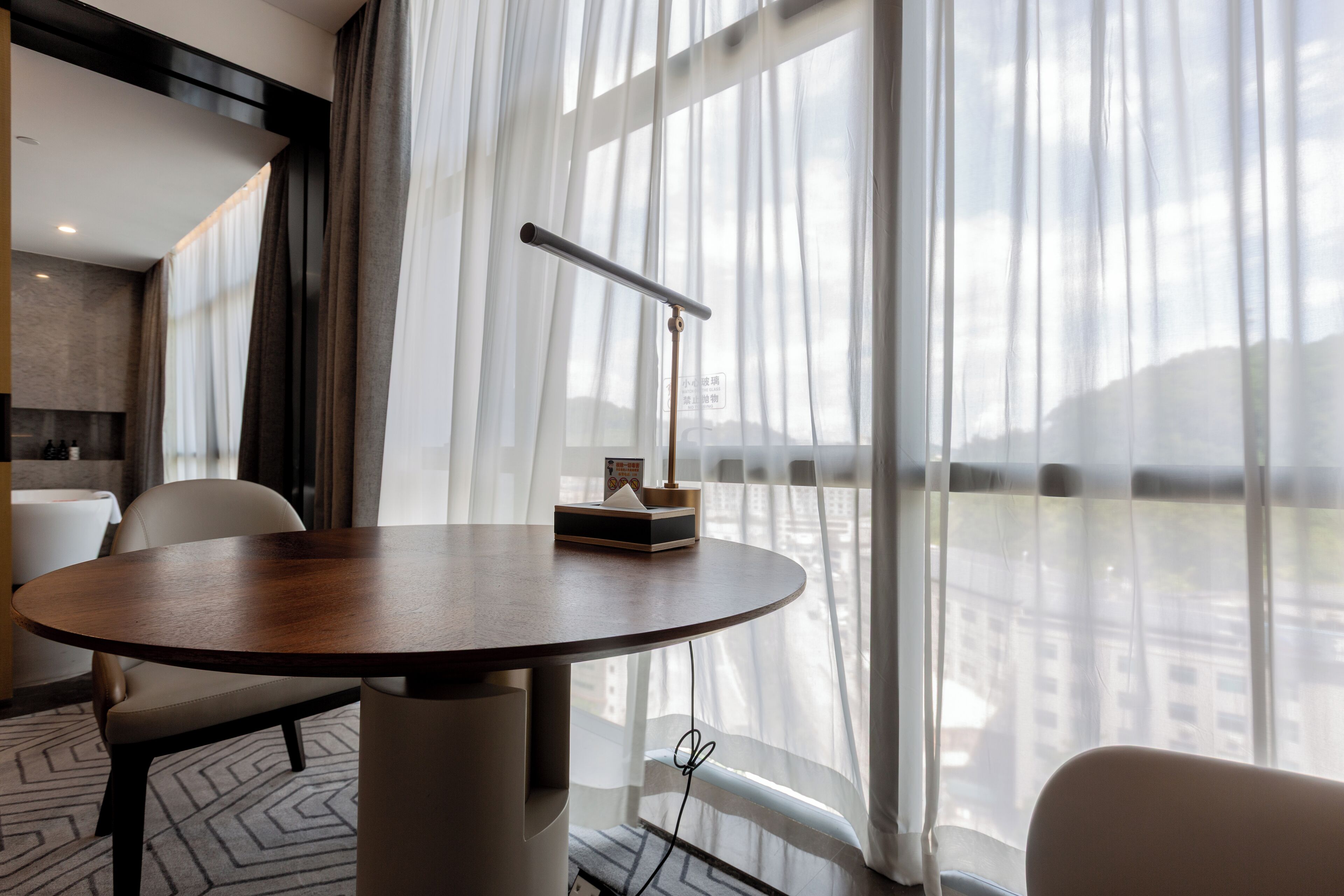 Une chambre d’hôtel moderne avec une table ronde, une chaise, une lampe et une vue sur la ville à travers des rideaux transparents.