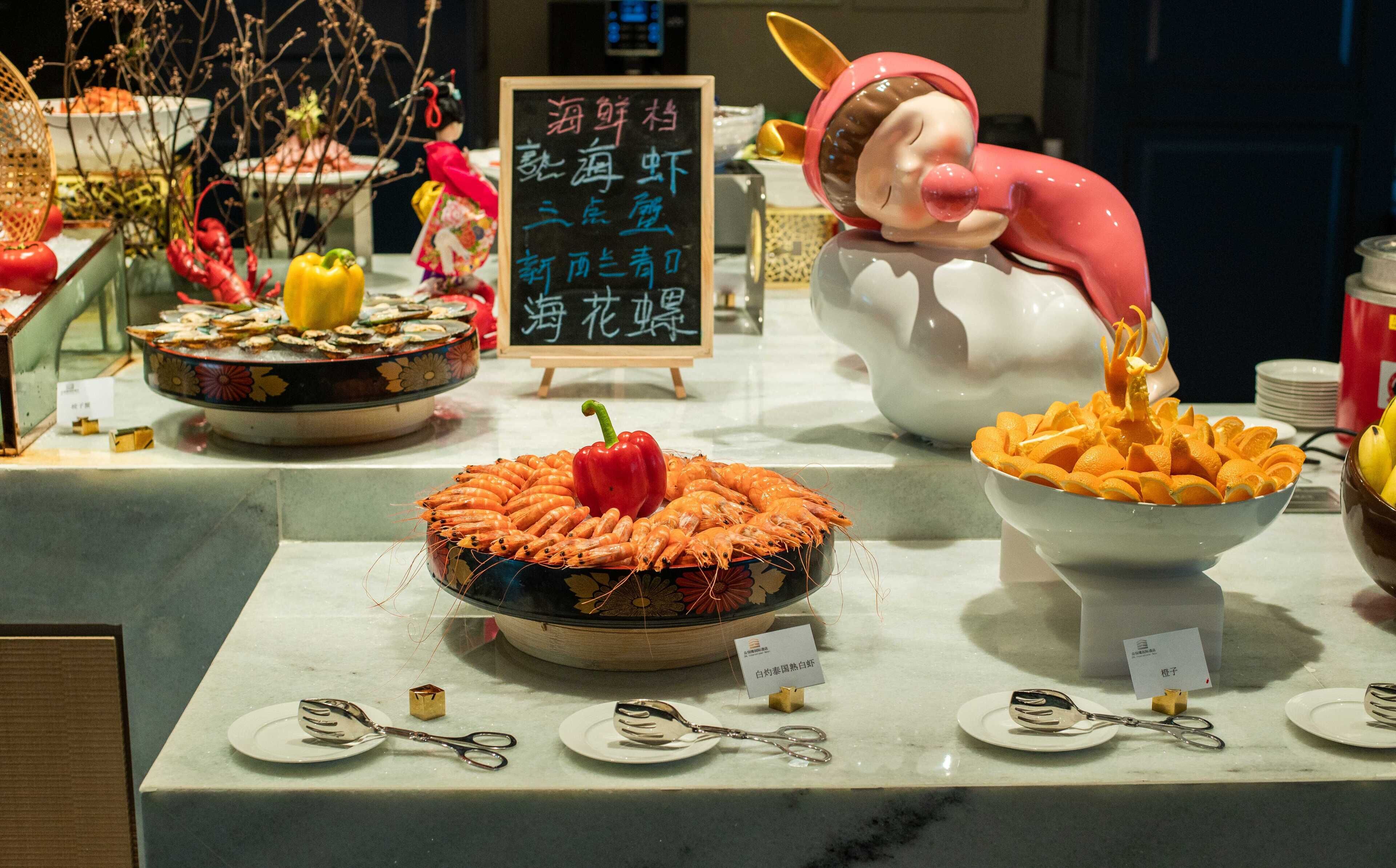 Une table de buffet proposant divers plats, notamment des crevettes et des tranches d’orange, ainsi qu’une figurine décorative.