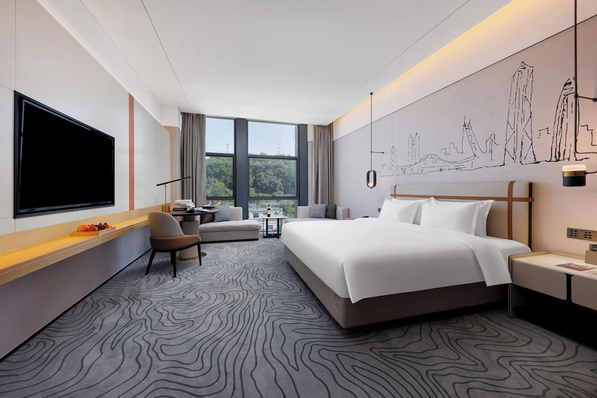 A modern hotel room with a large bed, a flat-screen TV, a sofa, a desk, and a cityscape mural on the wall.