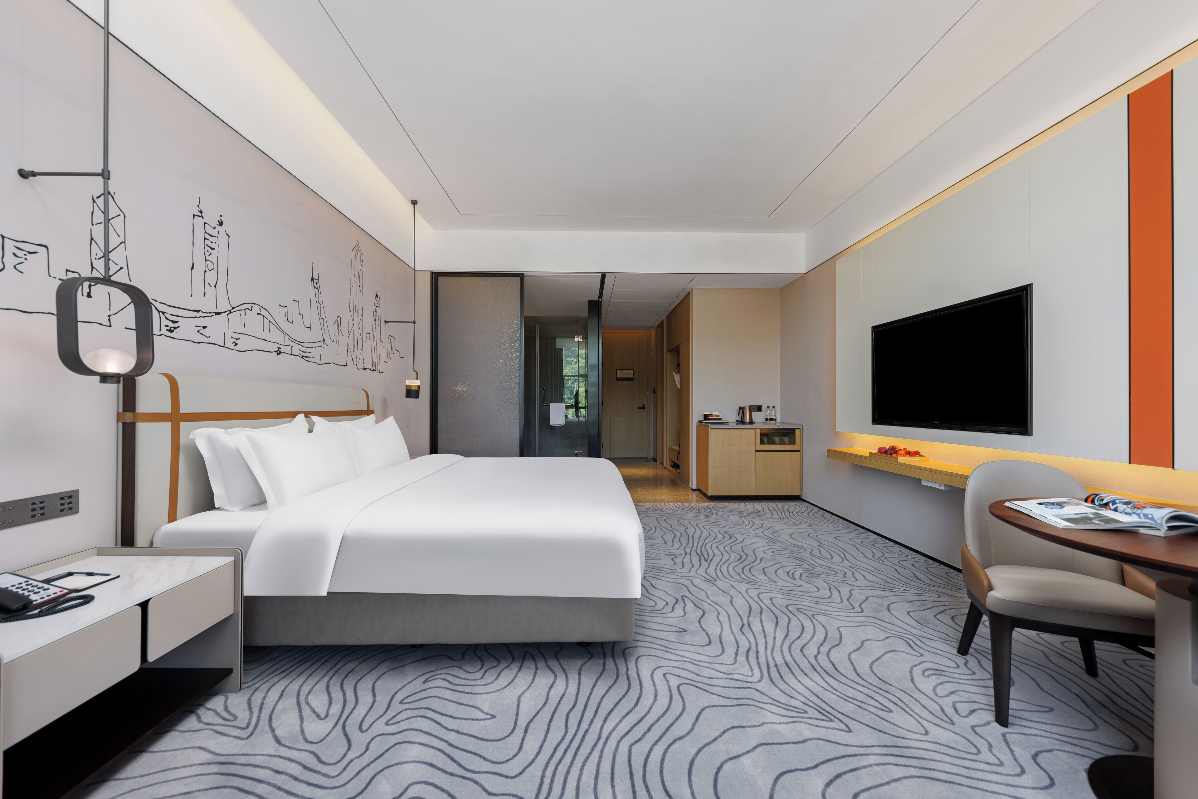 A modern hotel room with a large bed, a desk, a chair, a TV, and a kitchen area.