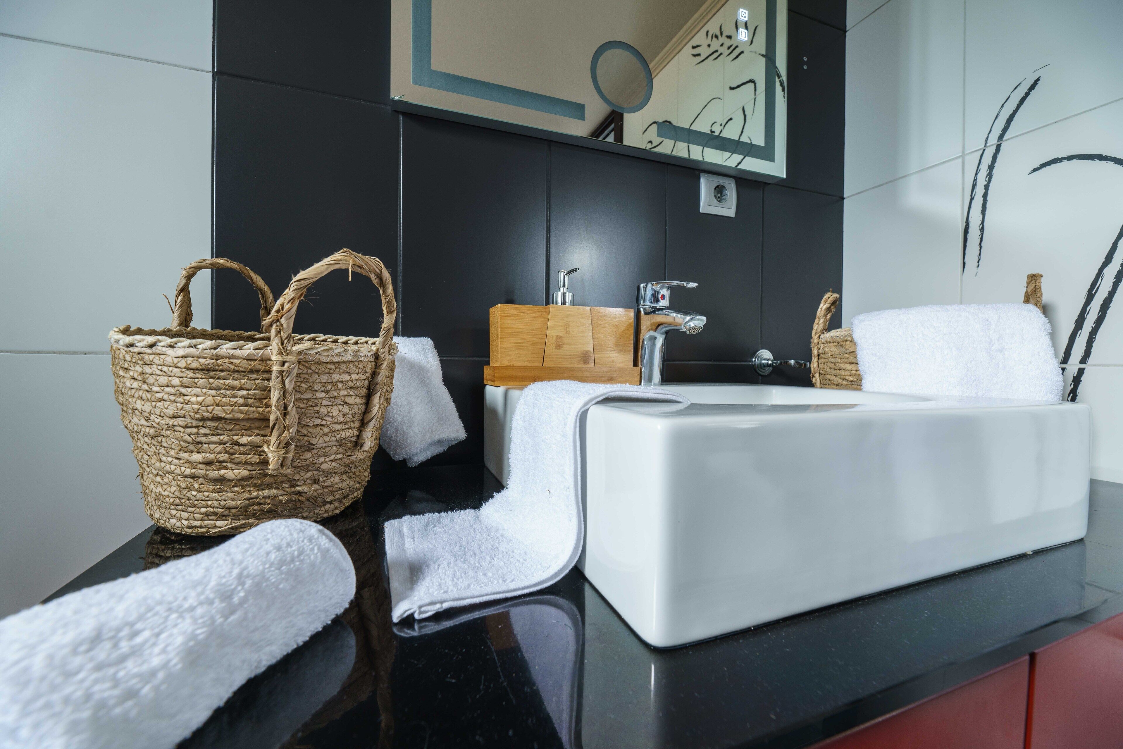 A modern bathroom with a black countertop, white sink, and a wooden vanity. There are two woven baskets and a white towel on the counter.