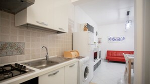 Cocina compacta con estufa a gas, lavamanos y lavadora, que da paso a una sala de estar con sofá rojo y una mesa de comedor.