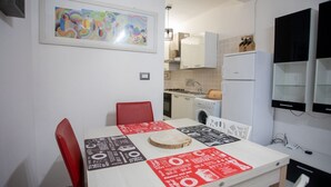 Una cocina pequeña y moderna con armarios blancos, un refrigerador y una mesa de comedor con sillas rojas. Hay un cuadro abstracto enmarcado en la pared sobre la mesa.
