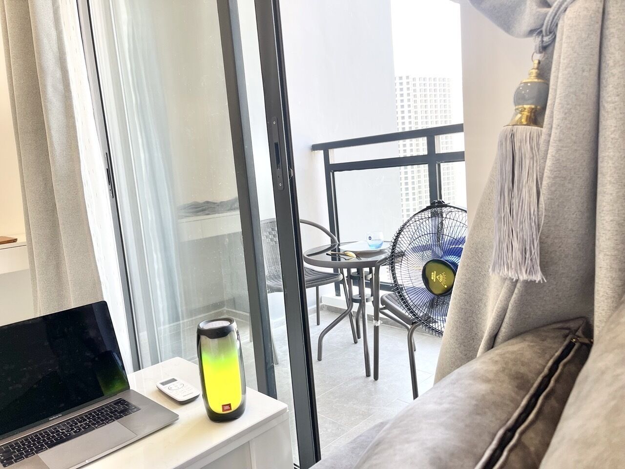 A modern room with a laptop on a desk, a standing heater, and a view of an outdoor balcony with chairs and a fan.