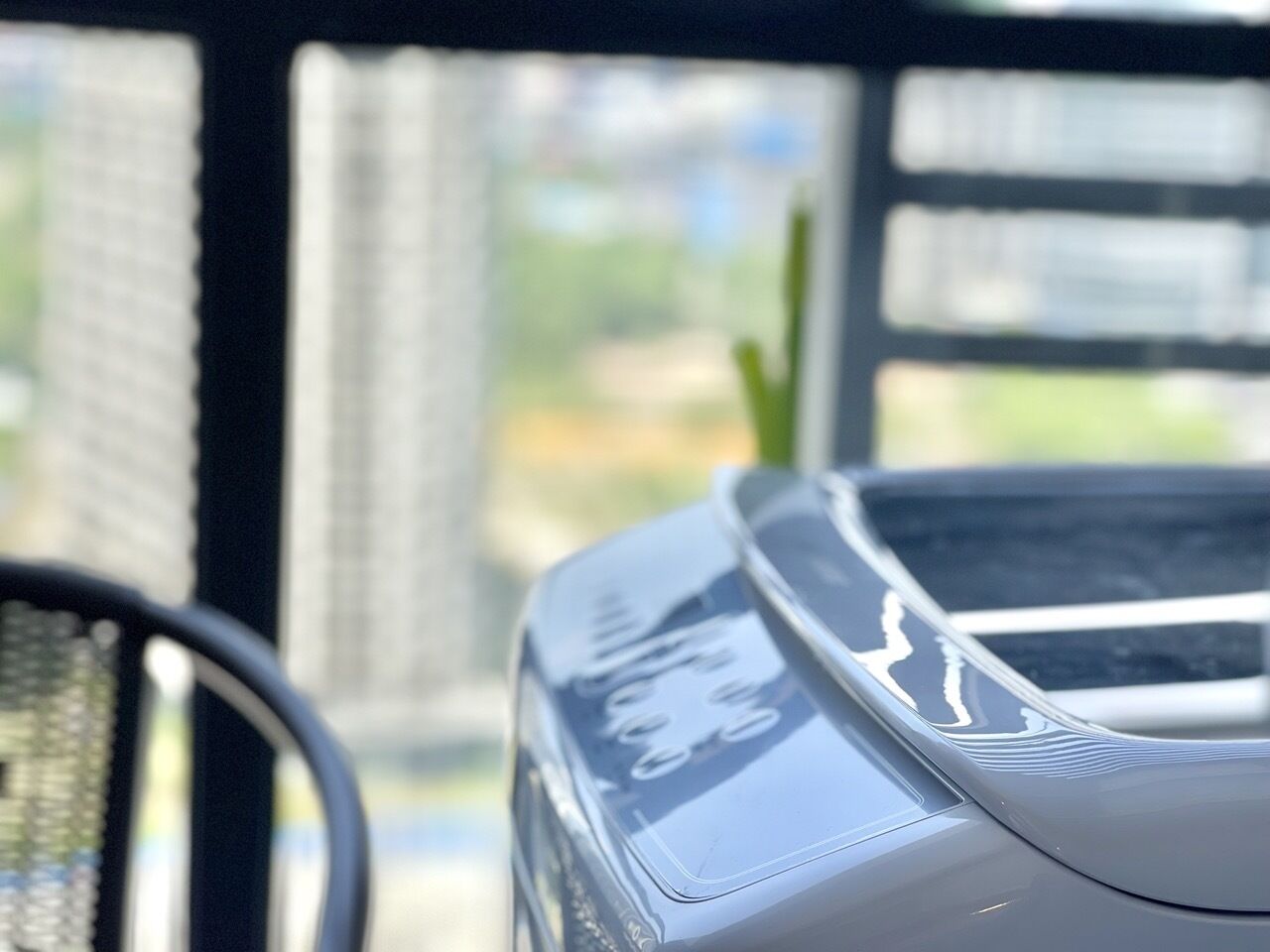 A close-up of a modern, sleek car with a reflective surface, parked near a window with a view of tall buildings and greenery.