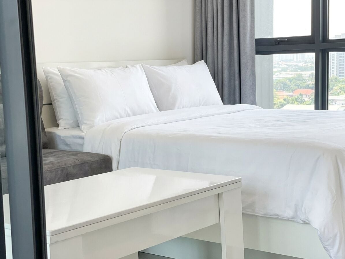 A modern hotel room with a bed, a bedside table, and a view of the city through large windows.