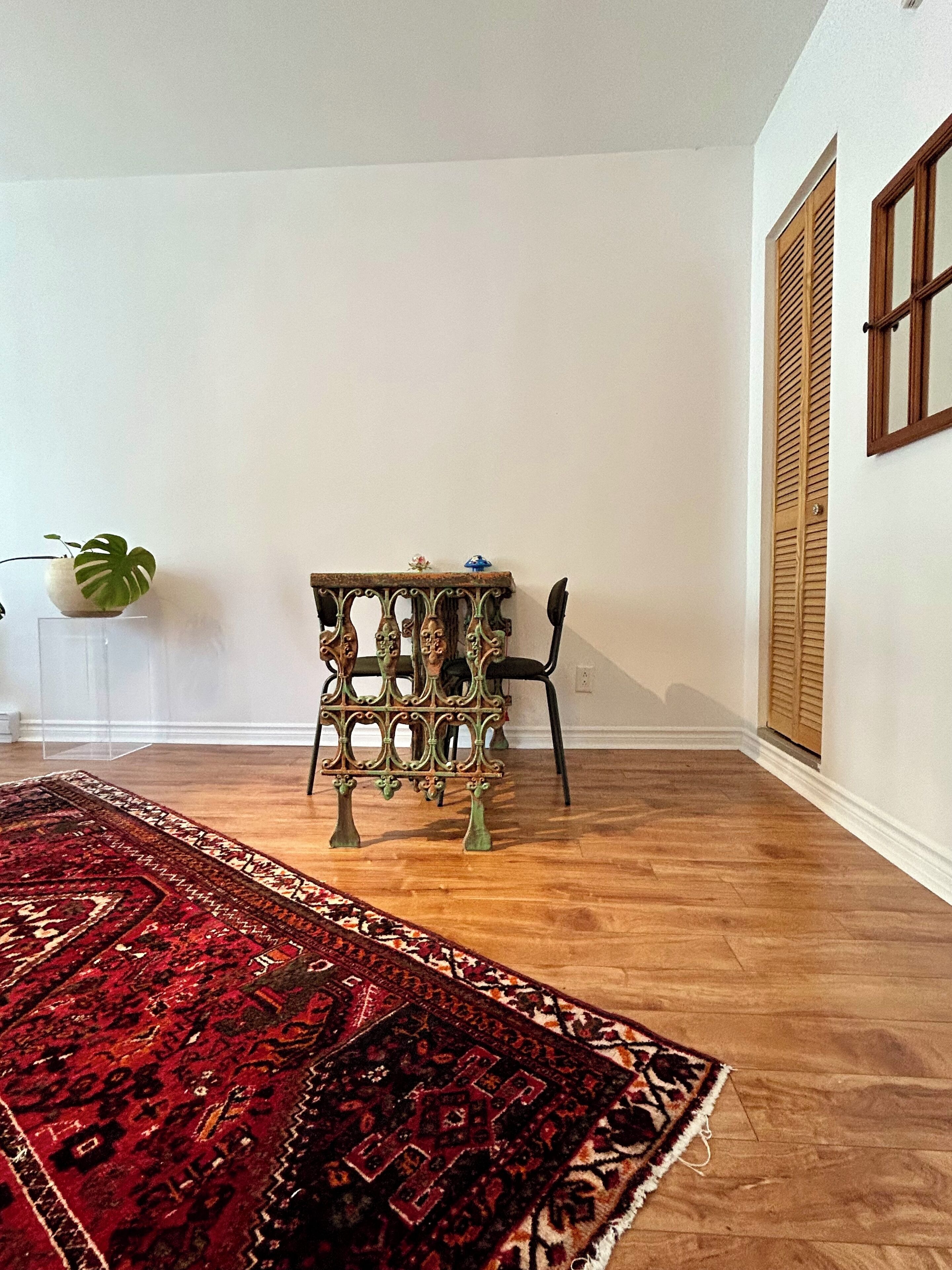 A room with a wooden floor, a red patterned rug, a metal table with a decorative top, and a potted plant.