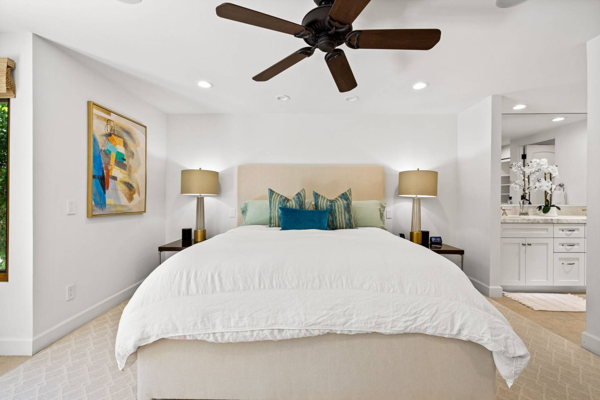 A modern bedroom with a large bed, two bedside tables with lamps, a ceiling fan, a painting on the wall, and a bathroom visible through an open door.