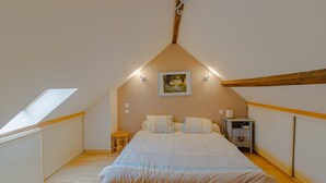 Un dormitorio con techo inclinado, una cama con cabecera de madera, una mesita de noche, un taburete pequeño y un cuadro en la pared.