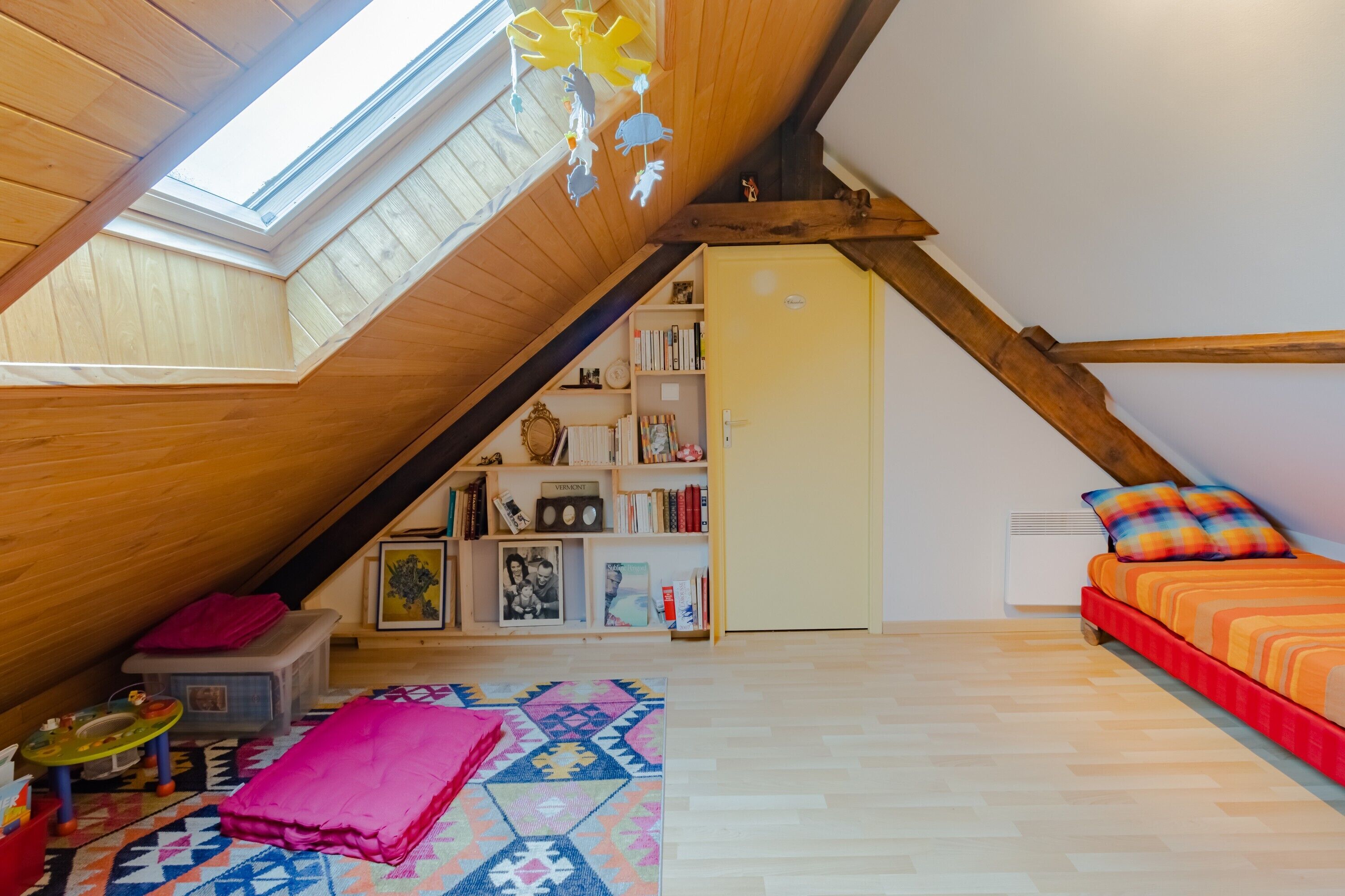 A room with a sloped ceiling, a skylight, a bookshelf, a bed with colorful bedding, and a patterned rug.