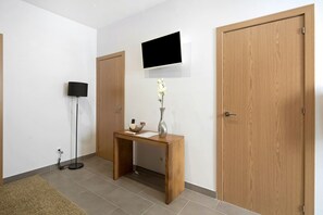 Un couloir avec un bureau en bois, un vase de fleurs, une lampe sur pied et un téléviseur à écran plat fixé au mur.