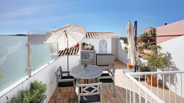 Una terrazza sul tetto con tavolo e sedie, un ombrellone bianco, piante in vaso e un muro decorato con piastrelle.