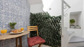 A corner with a patterned wall, a wooden chair, a table with a plate of food and drinks, and a green wall with plants.