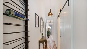 Un corridoio con pavimento decorato, un portariviste metallico con una bottiglia, un tavolo di legno con una pianta e un lampadario.