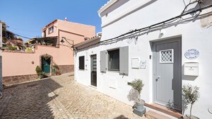 Un'angusta strada acciottolata con un edificio bianco, una porta grigia, piante in vaso e un cartello con la scritta 'Casa 16'.