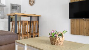 Una sala de estar moderna con una mesa de madera, una cesta de mimbre con flores, un televisor montado en la pared y una zona de cocina al fondo.