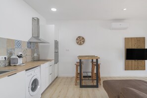 A modern kitchen with a washing machine, a wooden table with stools, a TV mounted on the wall, and a large window.