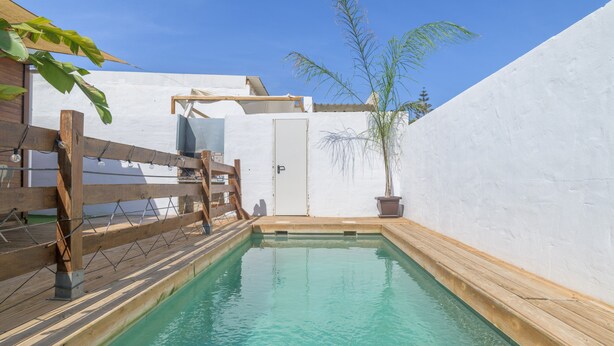 Una pequeña piscina rectangular con terraza de madera, rodeada de paredes blancas y una puerta que da a un área cubierta.