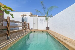 Una pequeña piscina rectangular con terraza de madera, rodeada de paredes blancas y una puerta que da a un área cubierta.