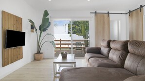 Una sala de estar moderna con un televisor de pantalla plana montado en un panel de madera, un sofá de cuero marrón, una mesa de centro blanca, una planta en maceta y un muro de vidrio corredizo que da a un patio exterior.
