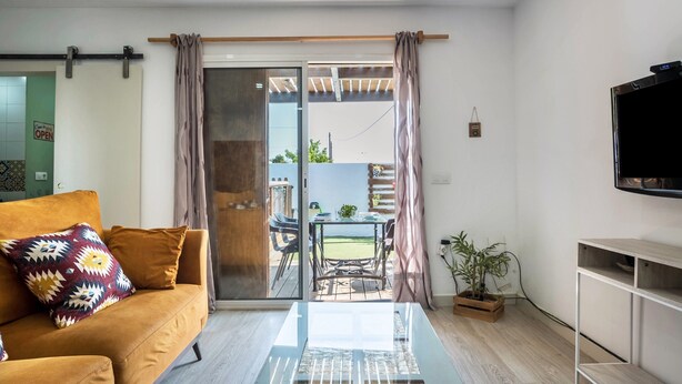 A living room with a sofa, a glass coffee table, a TV, a sliding glass door leading to an outdoor area with a table and chairs, and a potted plant.