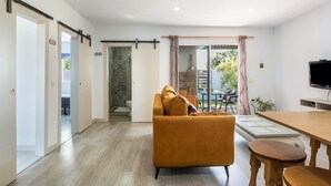 A modern living room with a sofa, a dining table, and a TV. There is a sliding door leading to a balcony with a view of greenery.