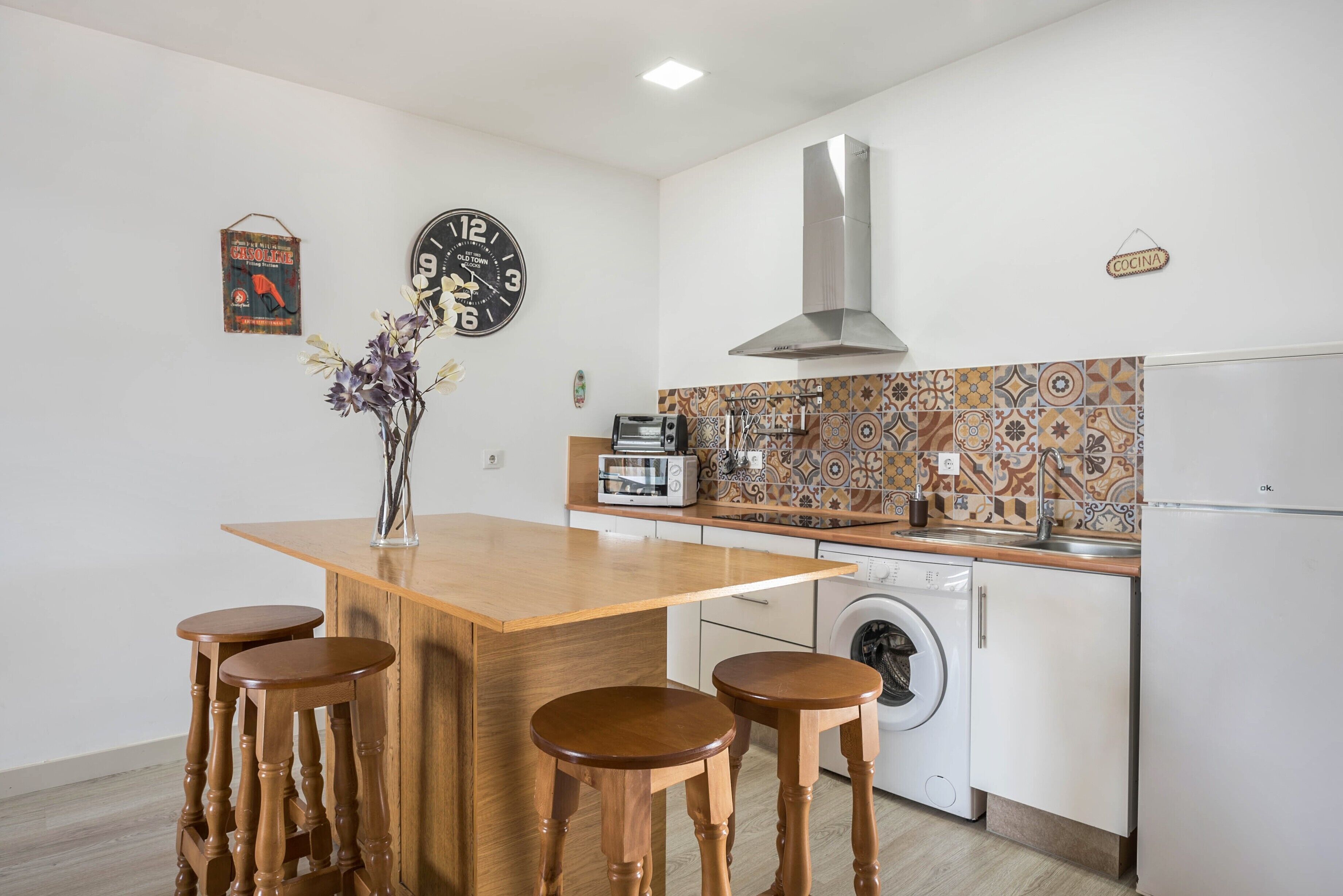 A compact kitchen with a central island, bar stools, a washing machine, and a clock on the wall.
