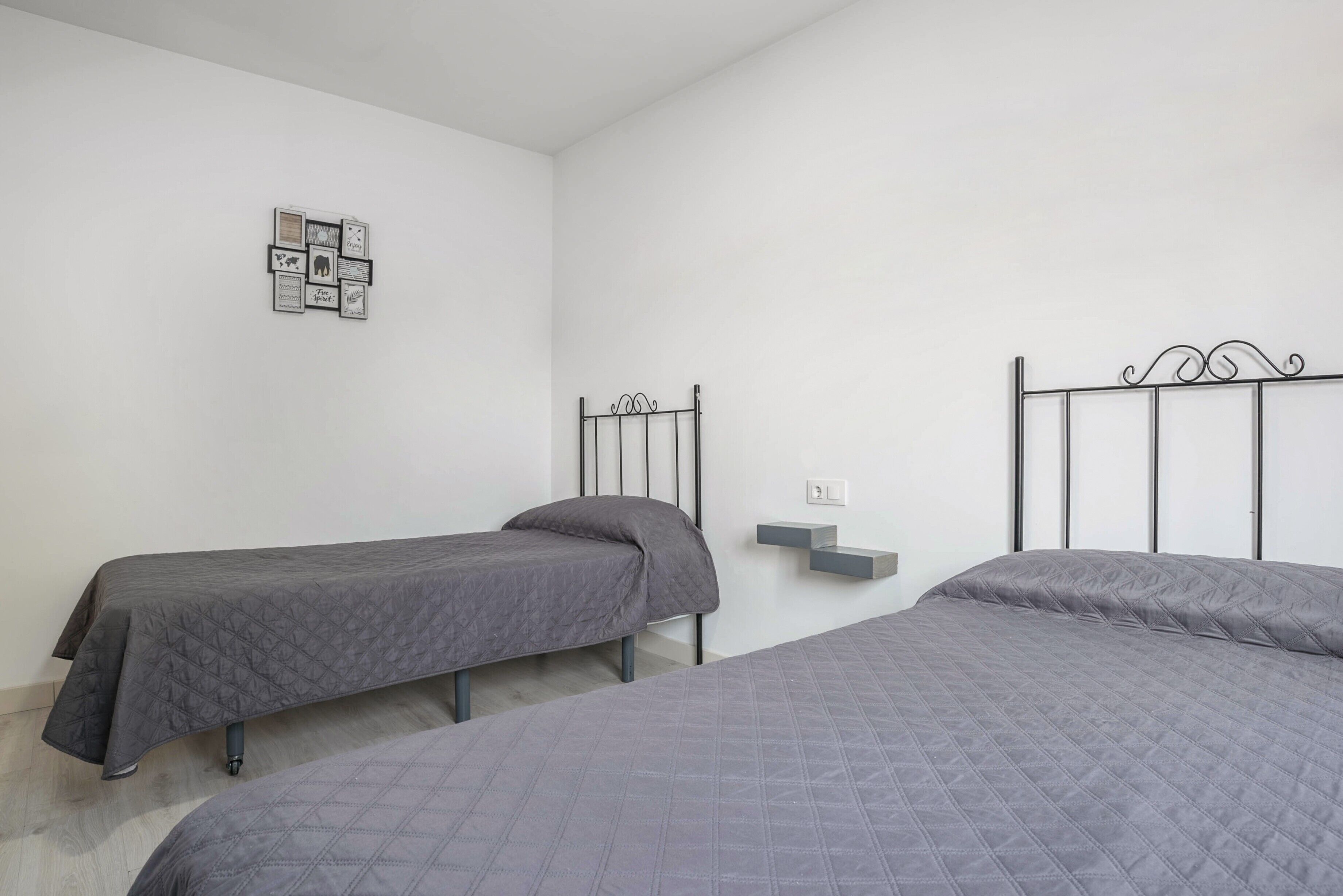 A bedroom with two beds, a wall-mounted shelf, and a framed picture collection.