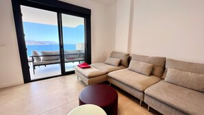 En stue med en sofa, en rød puf og et rundt bord. Der er en skydedør, der fører ud til en balkon med havudsigt.