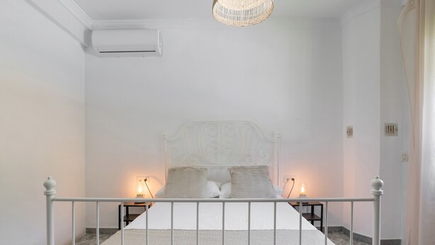 A bedroom with a white bed, two bedside tables with lamps, a white air conditioning unit, and a textured rug. - Chalet 'Casa El Molino' with Mountain View, Wi-Fi and Air Conditioning (Montecorto)