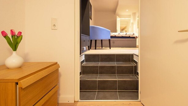 A hallway leading to a bedroom with a bed, a blue chair, and a white vase with flowers on a wooden table. - TOP SPOT- KREUZBURG CENTRAL THE PLACE TO BE ! (Berlin)