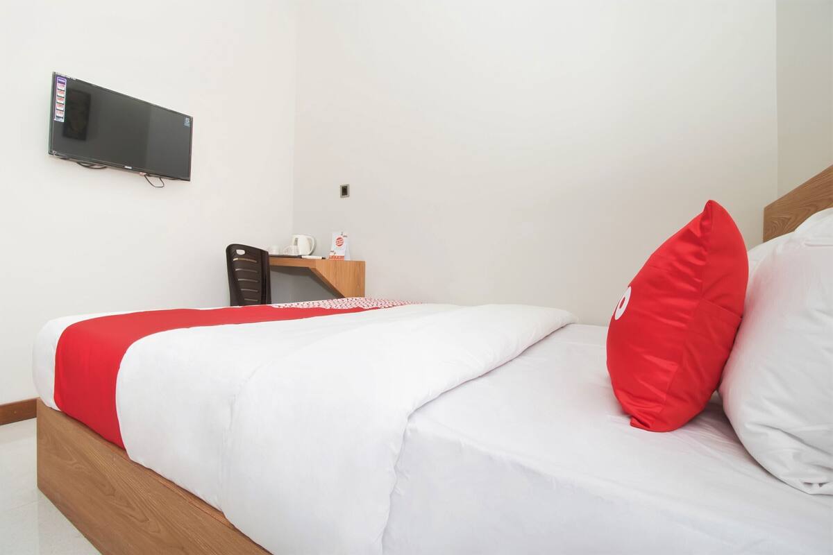 Un lit bien fait, recouvert d’une couverture rouge et blanche, avec un oreiller rouge et un oreiller blanc. Un téléviseur à écran plat est fixé au mur au-dessus du lit.