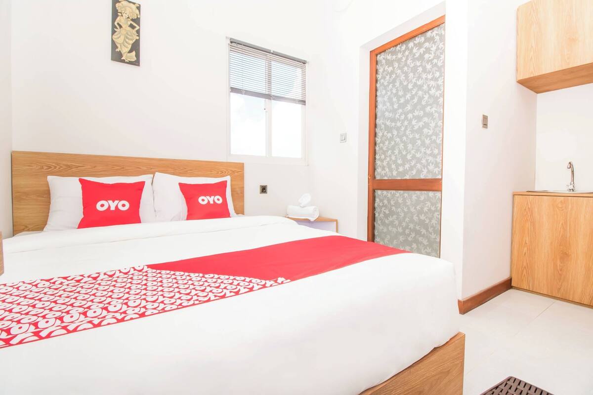 A hotel room with a bed, a window with blinds, a wooden headboard, a bedside table with a lamp, and a wardrobe.