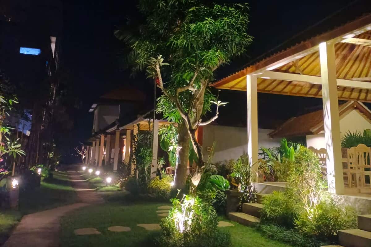 Vista noturna de um hotel com um caminho bem iluminado, área externa coberta e vegetação exuberante.