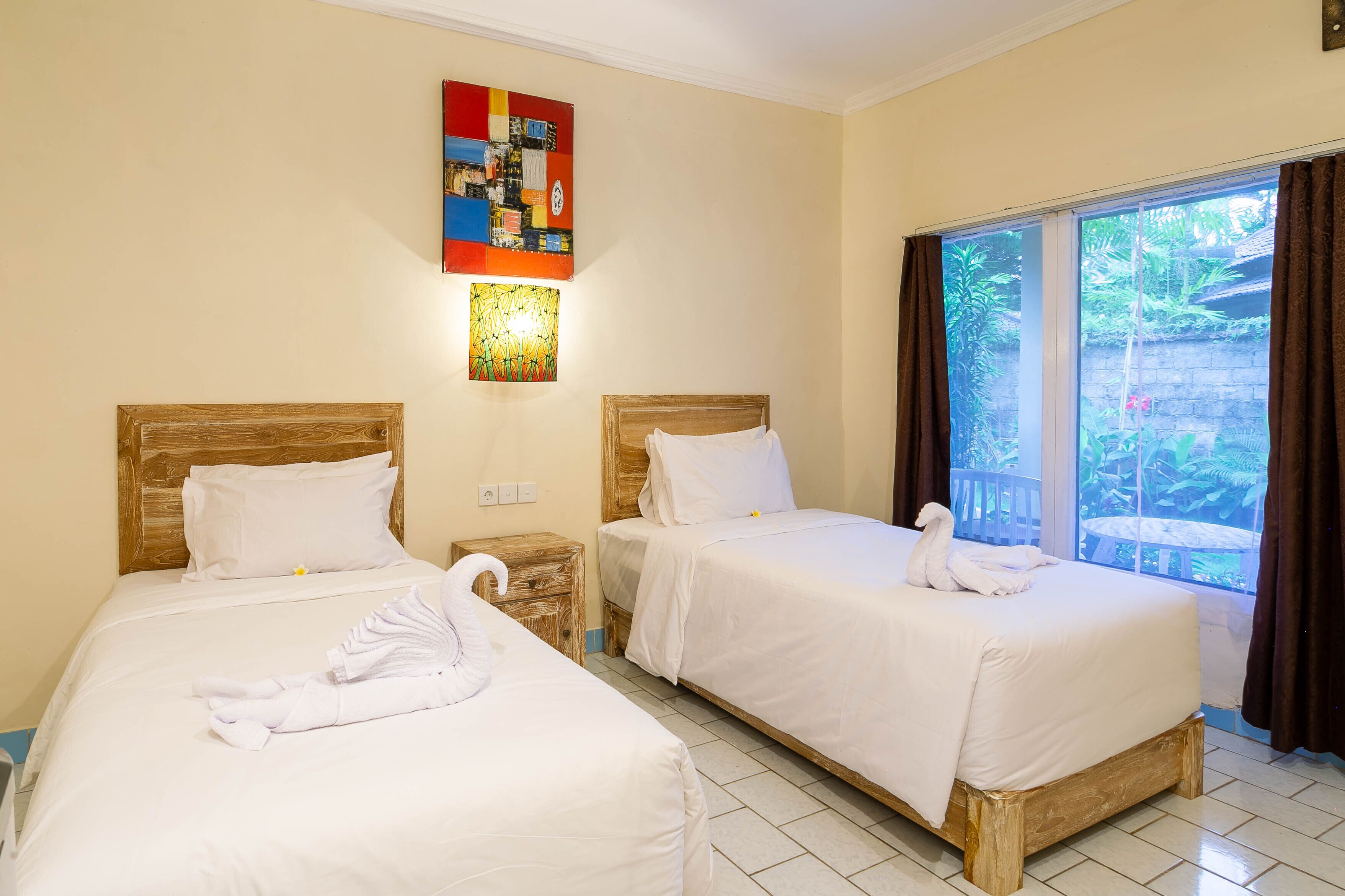 A hotel room with two beds, each with white linens and a wooden headboard. There is a painting on the wall above the beds and a window with curtains.
