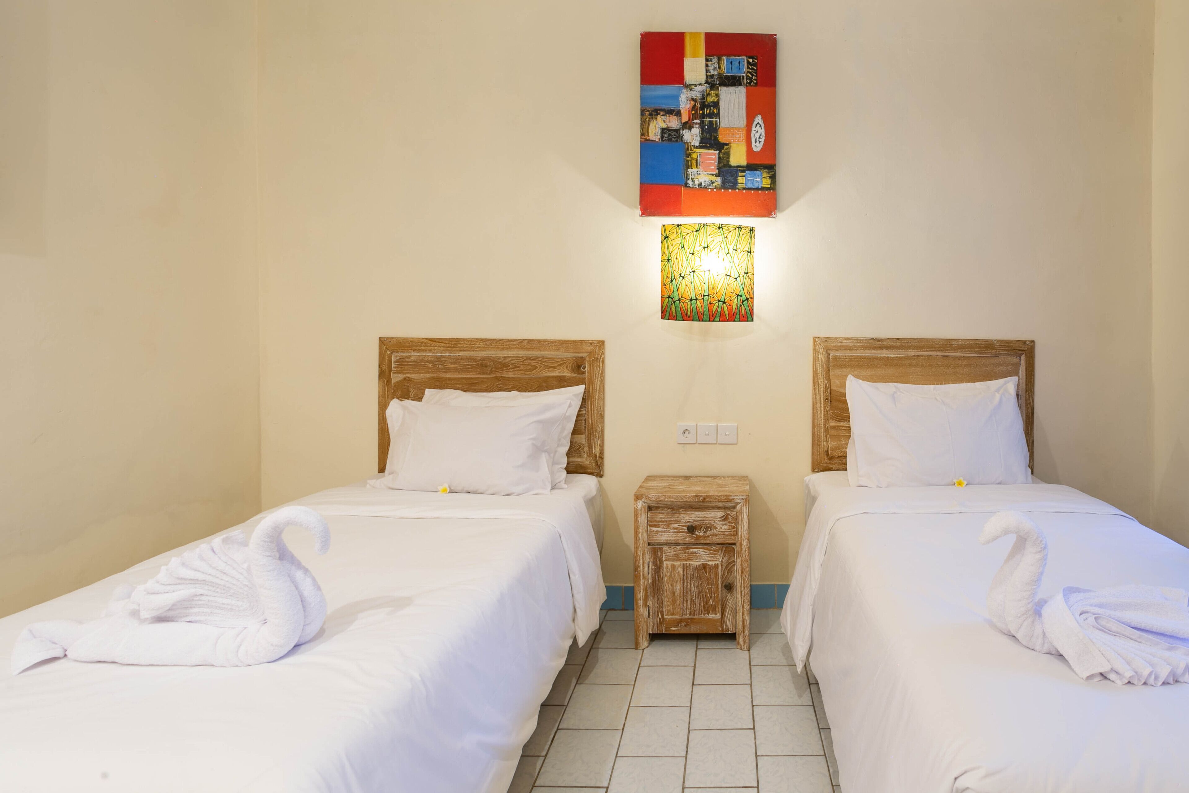 Habitación de hotel con dos camas individuales, cada una con cabecero de madera y ropa de cama blanca. Hay una mesita de noche de madera entre las camas, y una obra de arte colorida se exhibe sobre la mesita.
