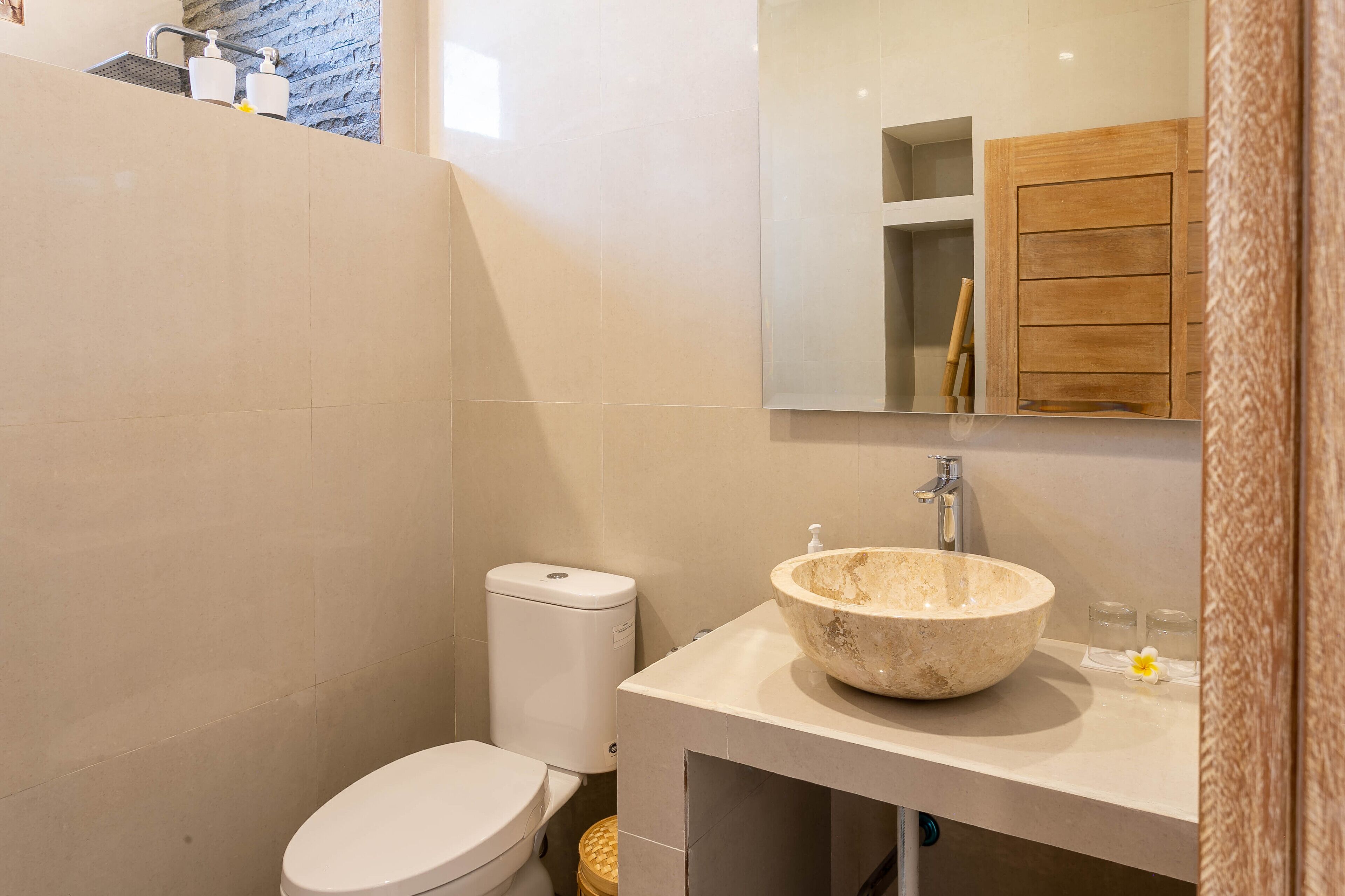 Un baño moderno con inodoro blanco, lavabo color beige con recipiente amplio y un mueble de madera sobre el lavabo.