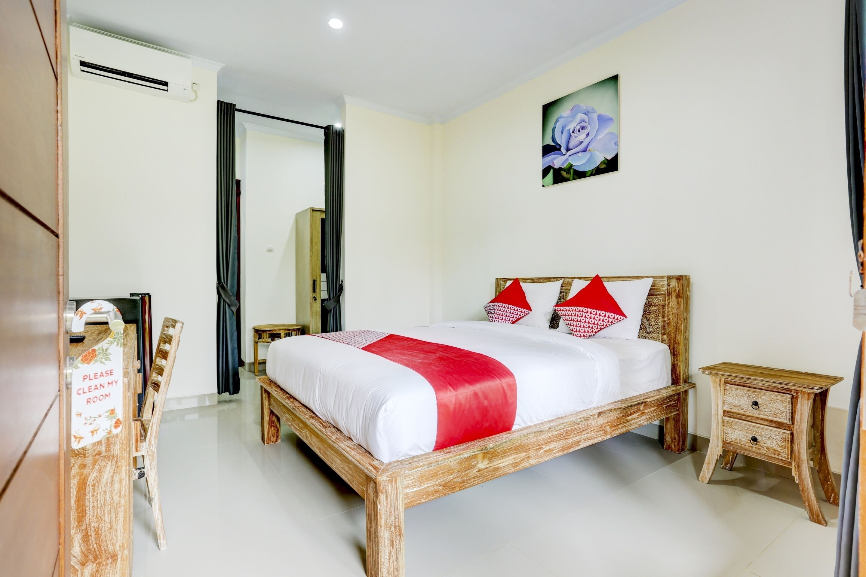 Une chambre d’hôtel avec un lit en bois, une couvre-lit rouge et blanche, une table de chevet en bois, une petite table, un miroir et un tableau représentant une fleur bleue accroché au mur.