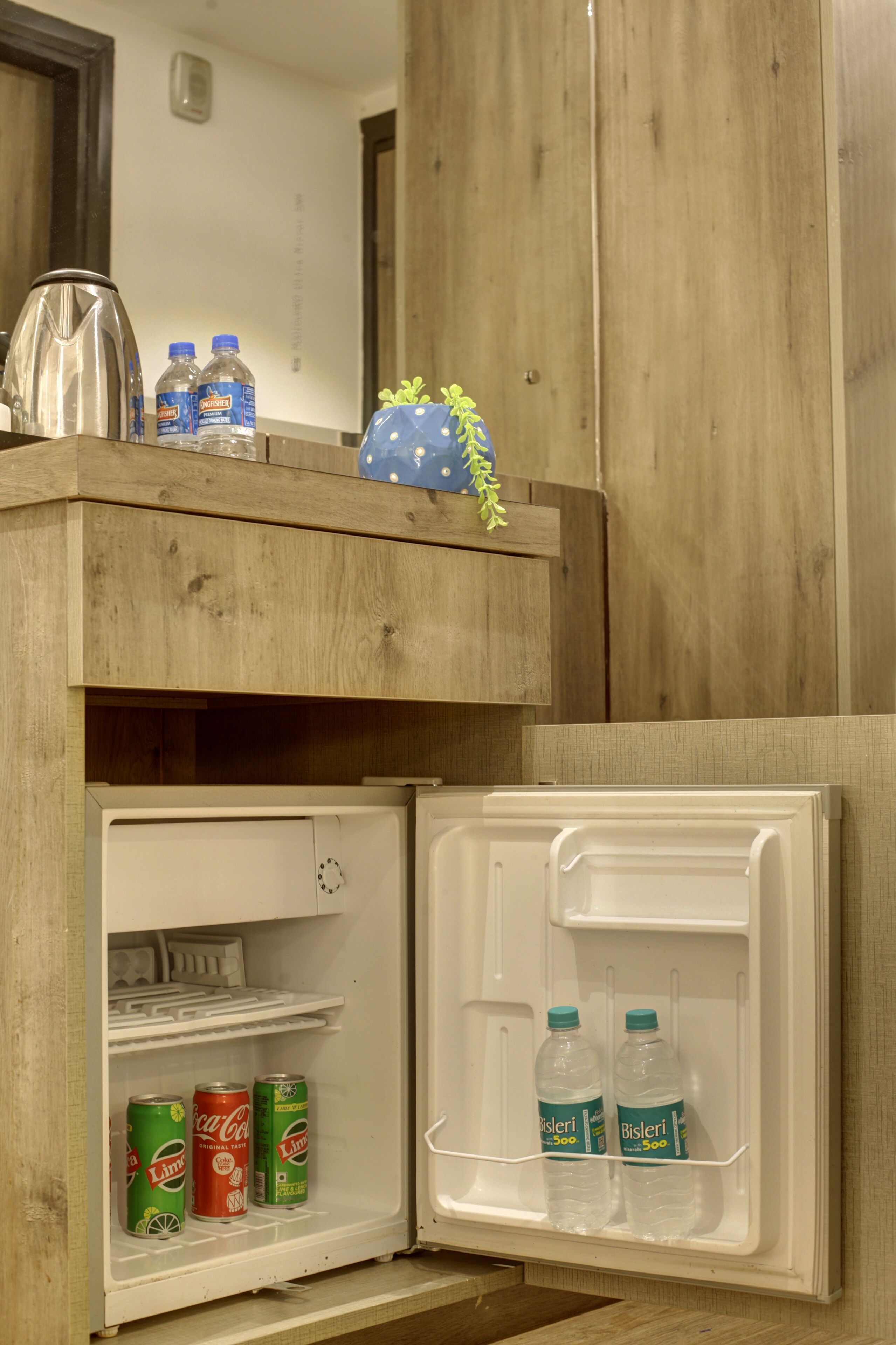 Eine Mini-Kühlbox mit einer Holzabdeckung, auf der ein Wasserkocher und Wasserflaschen stehen. Im Kühlschrank befinden sich Dosen Coca-Cola und Bisleri Wasser.