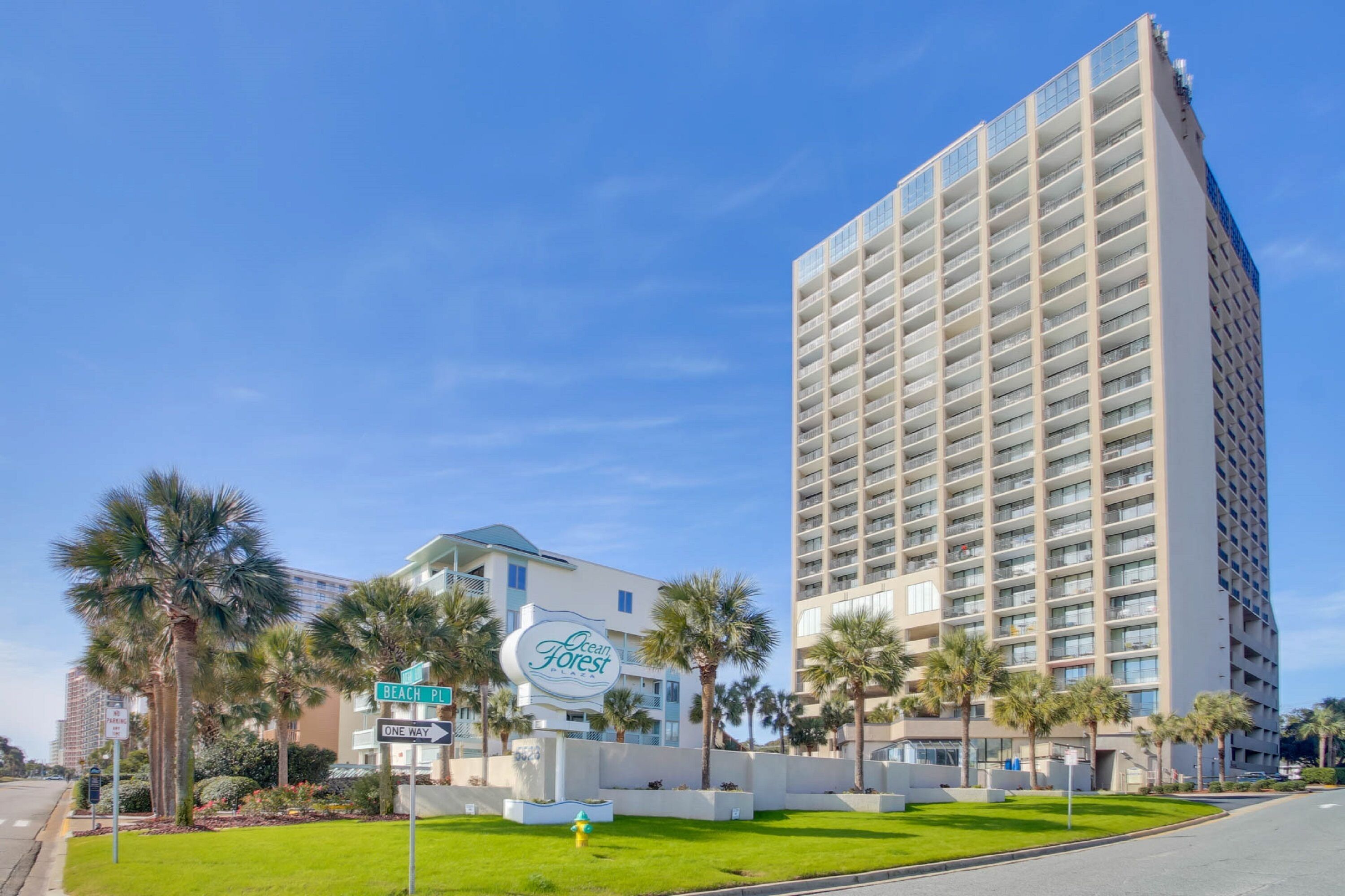 Een hoog hotel met een modern ontwerp, met een groot bord voor 'Ocean Forest' en een verzorgd gazon met palmbomen.