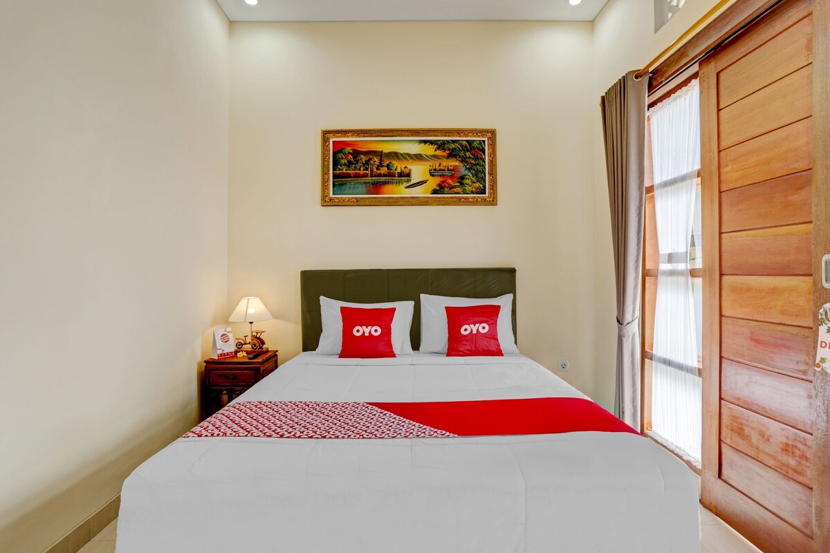 A hotel room with a bed, a nightstand, a lamp, a painting on the wall, and a wooden door.
